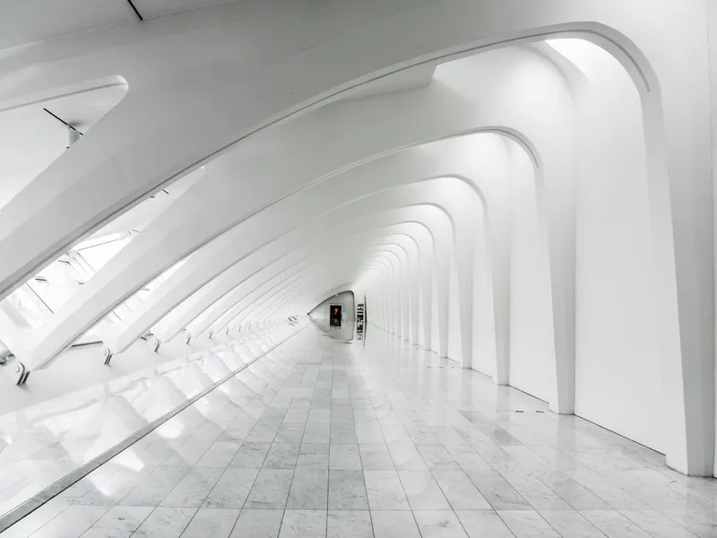 A white architectural corridor with repeating curved ribs and daylight across the floor.