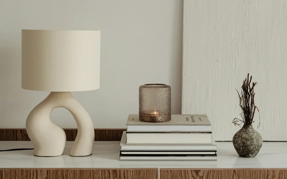 A sculptural cream table lamp, amber glass candle holder and small vase on a timber cabinet.